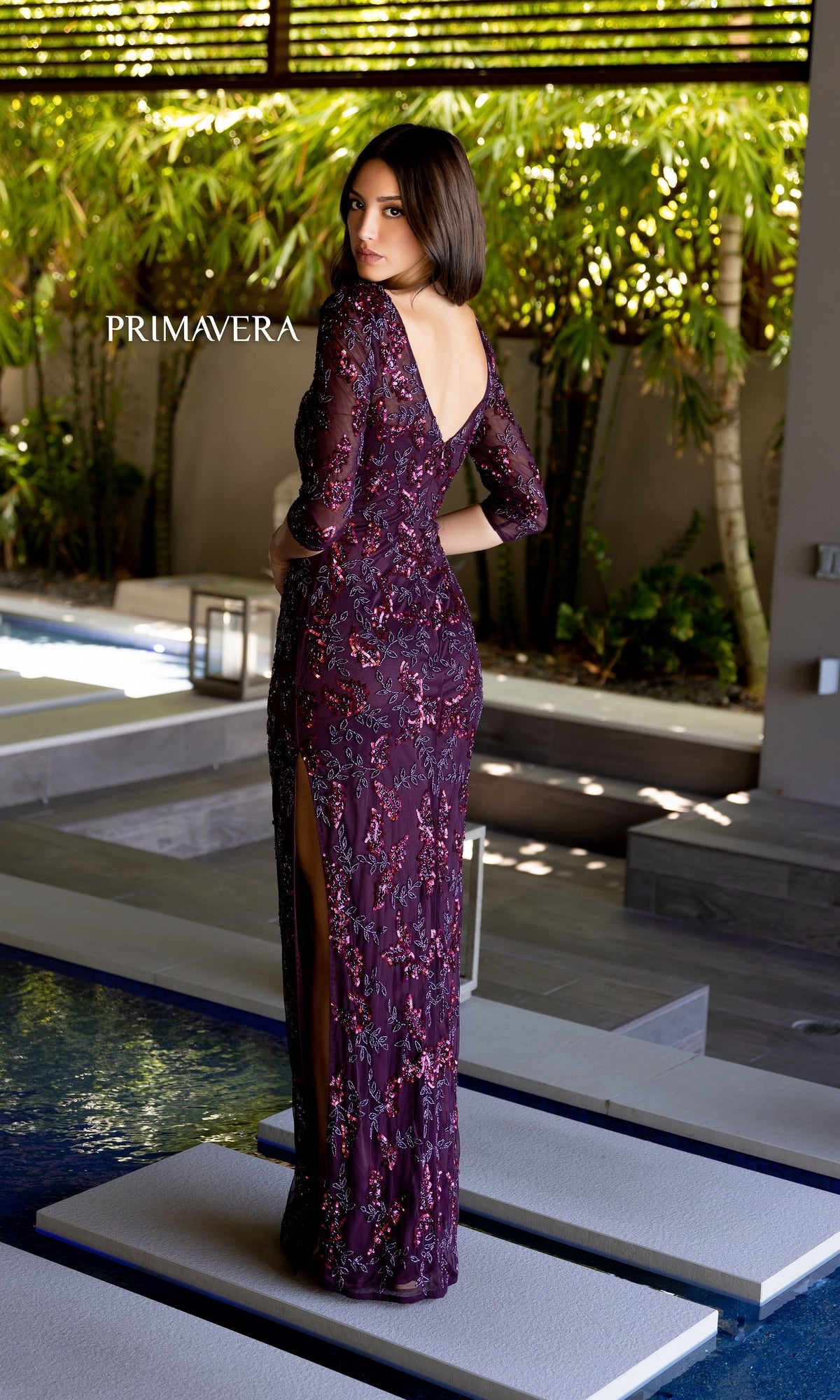 A woman in the Primavera 13116 Sleeved Long Beaded Formal Dress stands by a modern outdoor pool, wearing a deep purple gown with three-quarter sleeves and a back slit.